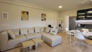 Ein modernes Wohnzimmer mit einem beigefarbenen Ecksofa, einem kleinen Holztisch und einem Essbereich mit weißem Tisch und Stühlen.