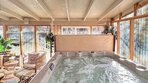 A hot tub in a room with wooden beams, wicker furniture, and large windows.