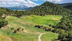 A golf course with a winding path, surrounded by dense forests and mountains in the background.
