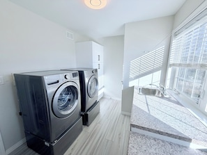 A laundry room with two front-loading washers and dryers, a window with blinds, and a white cabinet. - Chateau Sage Creek on the Lake, Hot Tub, Pool, Breathtaking sunsets, Nature path (Winnipeg)