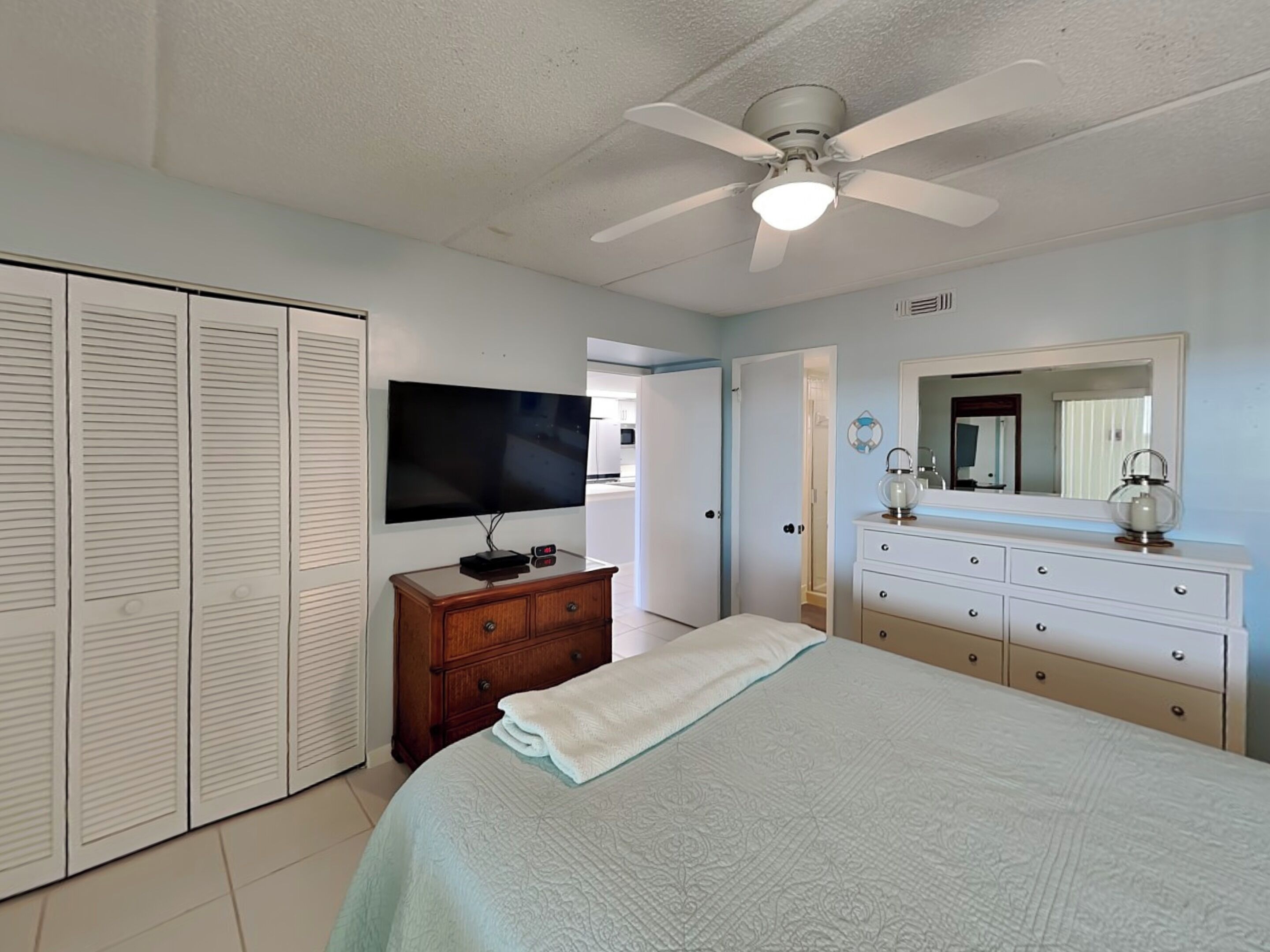Une chambre à coucher comprenant un lit, un téléviseur posé sur une commode en bois, un ventilateur de plafond, une commode, un miroir et une armoire.