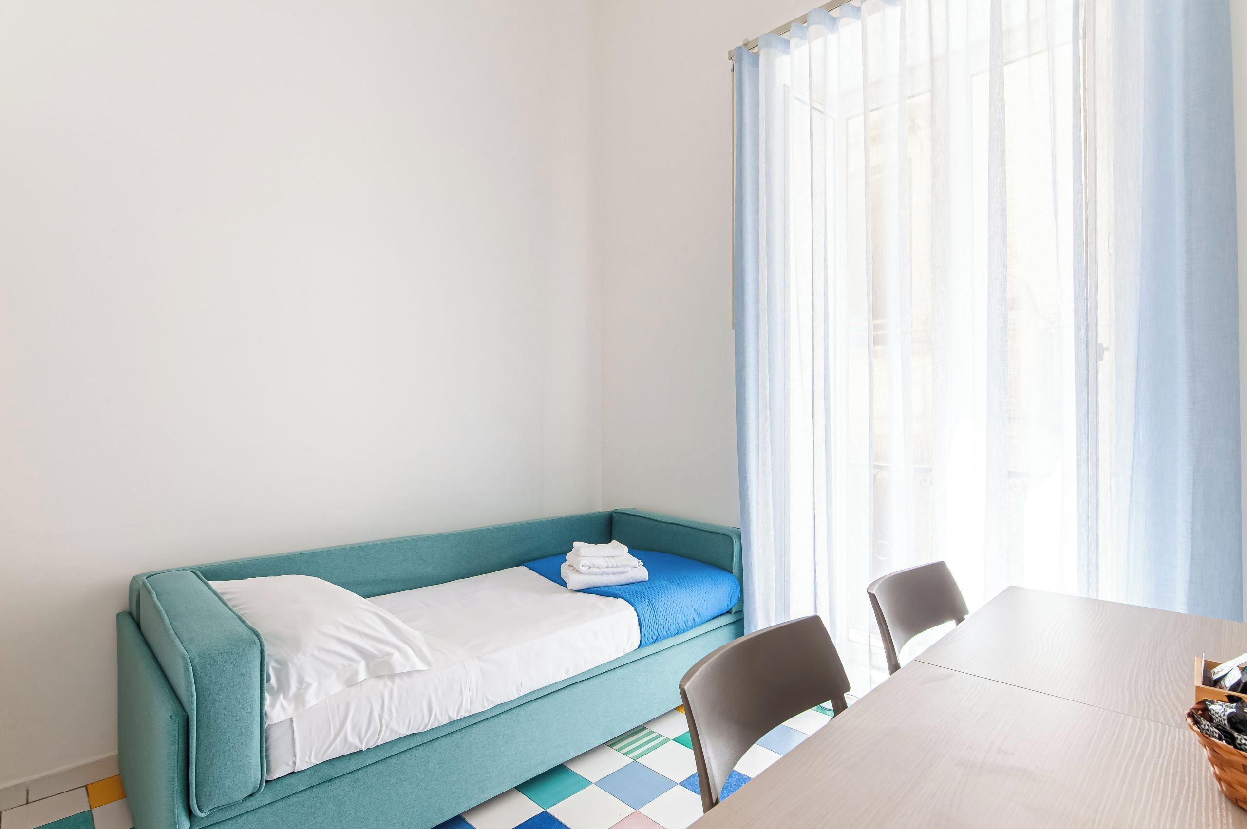 A single-bed room with a teal-colored bed, white bedding, a wooden table, and a chair.