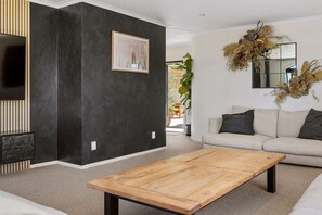 A modern living room with a dark accent wall, a large flat-screen TV, a beige sofa with dark cushions, a wooden coffee table, and a large mirror with dried flowers. - Rangatira Sunsets & Close to town (Taupō)