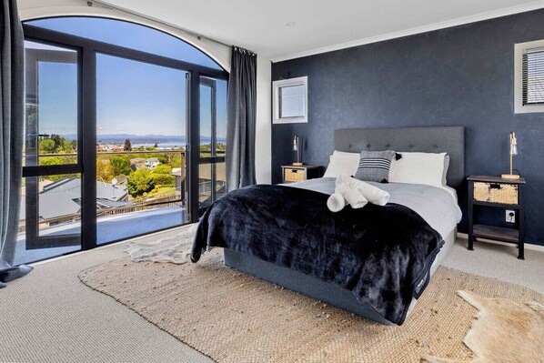 A bedroom with a large bed, a view through a glass door, a dark wall, and a nightstand with a lamp. - Rangatira Sunsets & Close to town (Taupō)