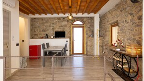 A room with a stone wall, wooden ceiling beams, a dining area with chairs and a table, a red appliance, and a clock on the wall.