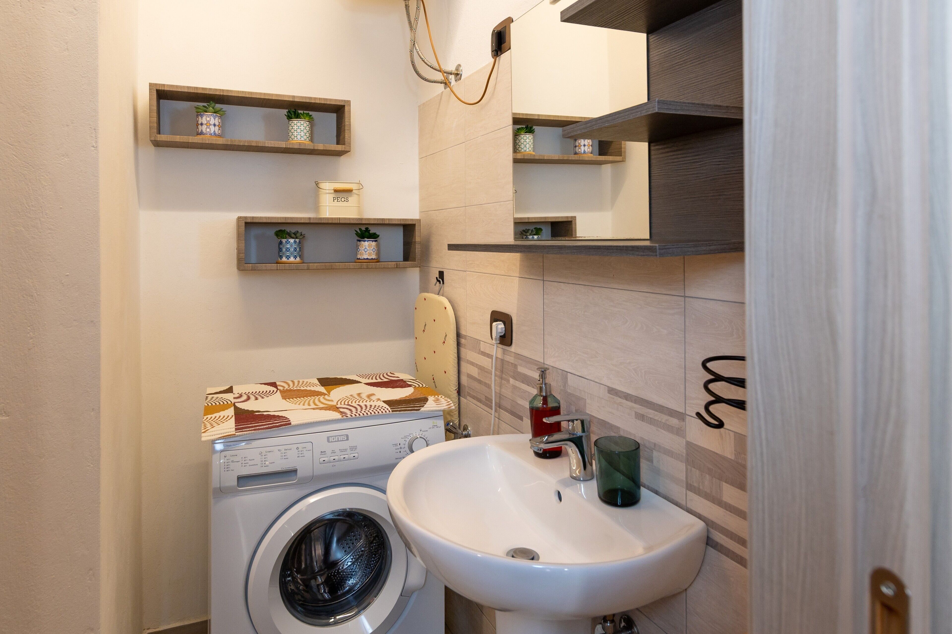 A compact bathroom with a washing machine, a sink, and wooden shelves with potted plants.
