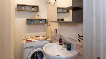 A compact bathroom with a washing machine, a sink, and wooden shelves with potted plants.