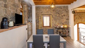 A stone-walled room with a wooden ceiling, a dining table with chairs, a television, and a small table with decorative items.