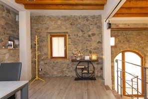 Una habitación con paredes de piedra, techo de madera, una mesa con una silla, un perchero, una pequeña mesa con adornos y una escalera con barandilla.