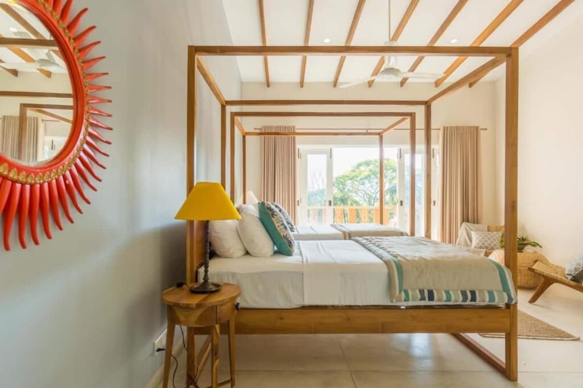 A room with a four-poster bed, a round mirror with a red frame, a small table with a lamp, and a view of the outdoors through large windows.