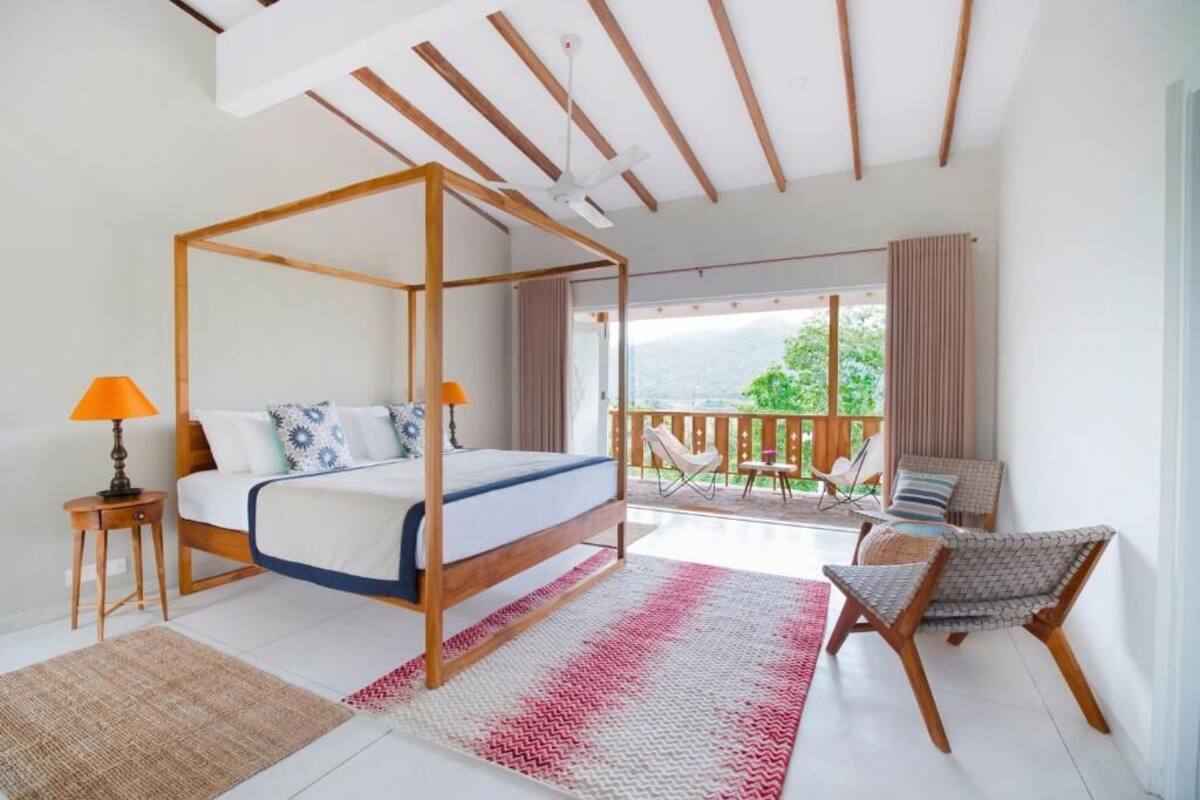 A bedroom with a four-poster bed, a balcony with chairs, and a view of greenery.