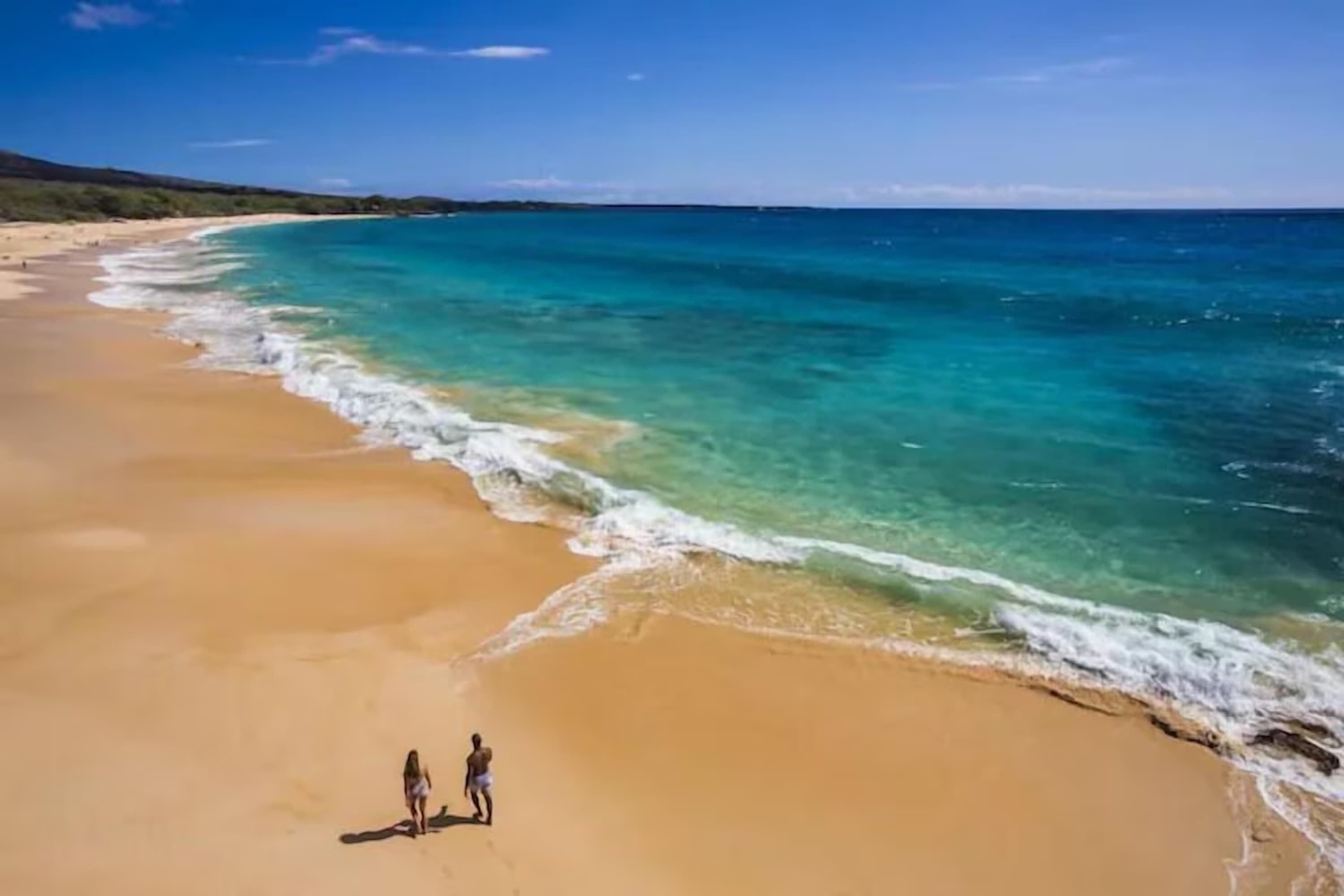 A wide sandy beach with clear turquoise waters and a few people walking along the shore.