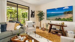 A living room with a large window, a sofa, a chair, a coffee table, a TV, and a painting of a beach. - Spacious Ground Floor Maui Retreat, Grand Champion (Wailea-Makena)