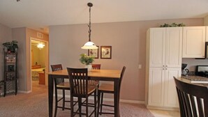 Een eetgedeelte met een houten tafel en stoelen, een hanglamp en een keuken met witte kastjes.