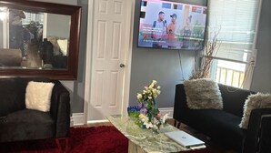 Ein Wohnzimmer mit rotem Shaggy-Teppich, zwei schwarzen Sofas mit weißen und braunen Kusselfern, einem Glastisch mit Blumenvase und einem an der Wand befestigten Flachbildfernseher.