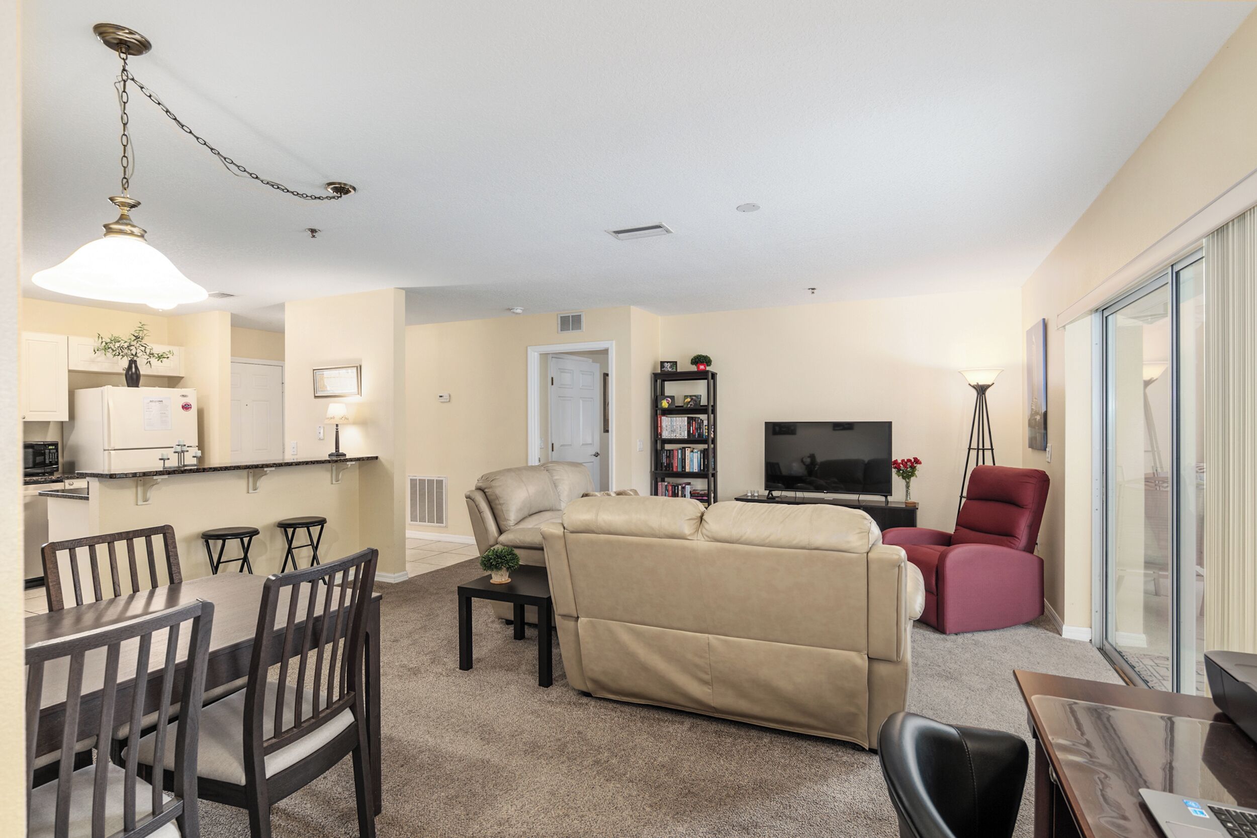 A living room with a sofa, a dining area with chairs, a kitchen with a refrigerator, and a TV in the background.