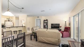 A living room with a sofa, a dining area with chairs, a kitchen with a refrigerator, and a TV in the background.