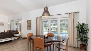Un área de comedor con una mesa redonda y sillas, una lámpara de techo y vistas a una piscina a través de amplias ventanas.