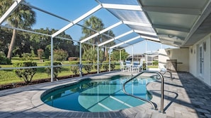 Una piscina exterior cubierta con zona de patio, una parrilla y vistas a un jardín ajardinado.