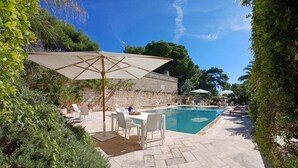 A poolside area with a table and chairs, surrounded by greenery and a stone wall.