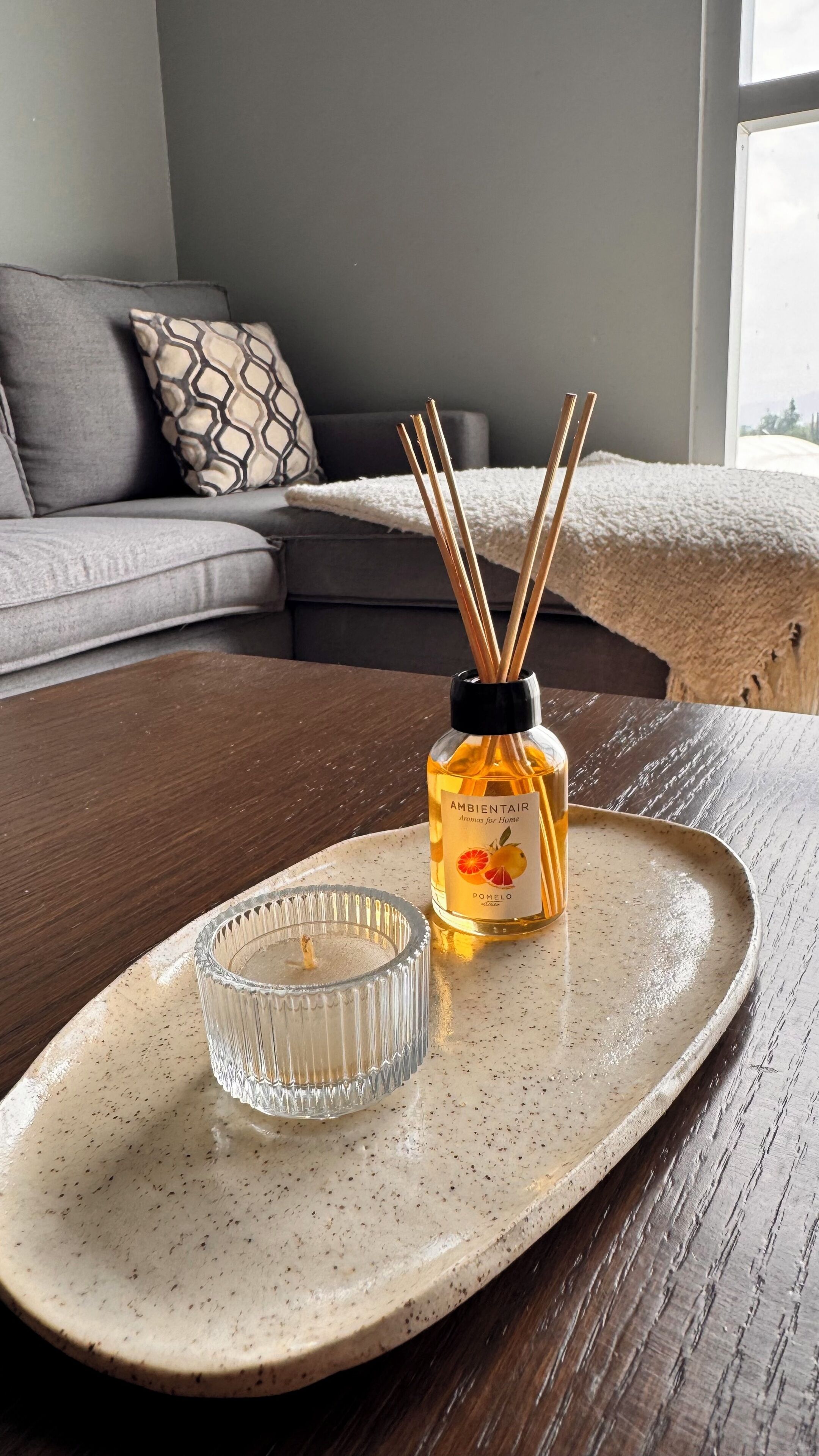 A ceramic tray with a glass candle holder and a bottle of AmbientaIR on a wooden table.