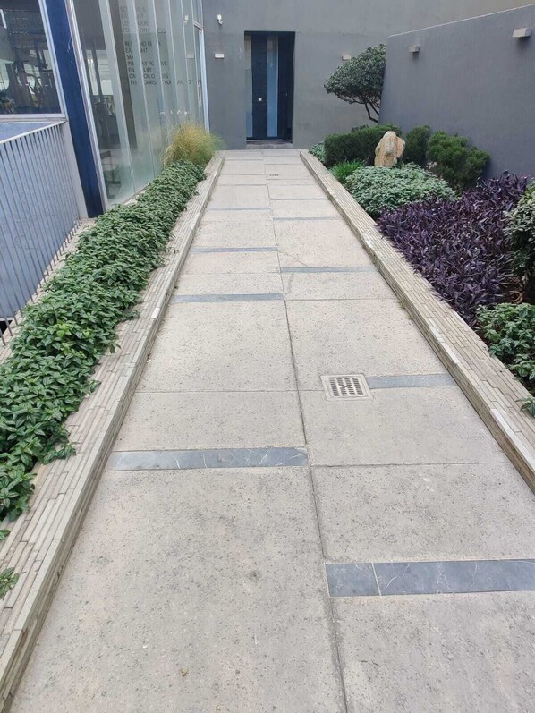 Een pad dat leidt naar een gebouwtoegang, omlijst door groen en paars plantenleven, met een stenen vloertegels en een metalen rooster.
