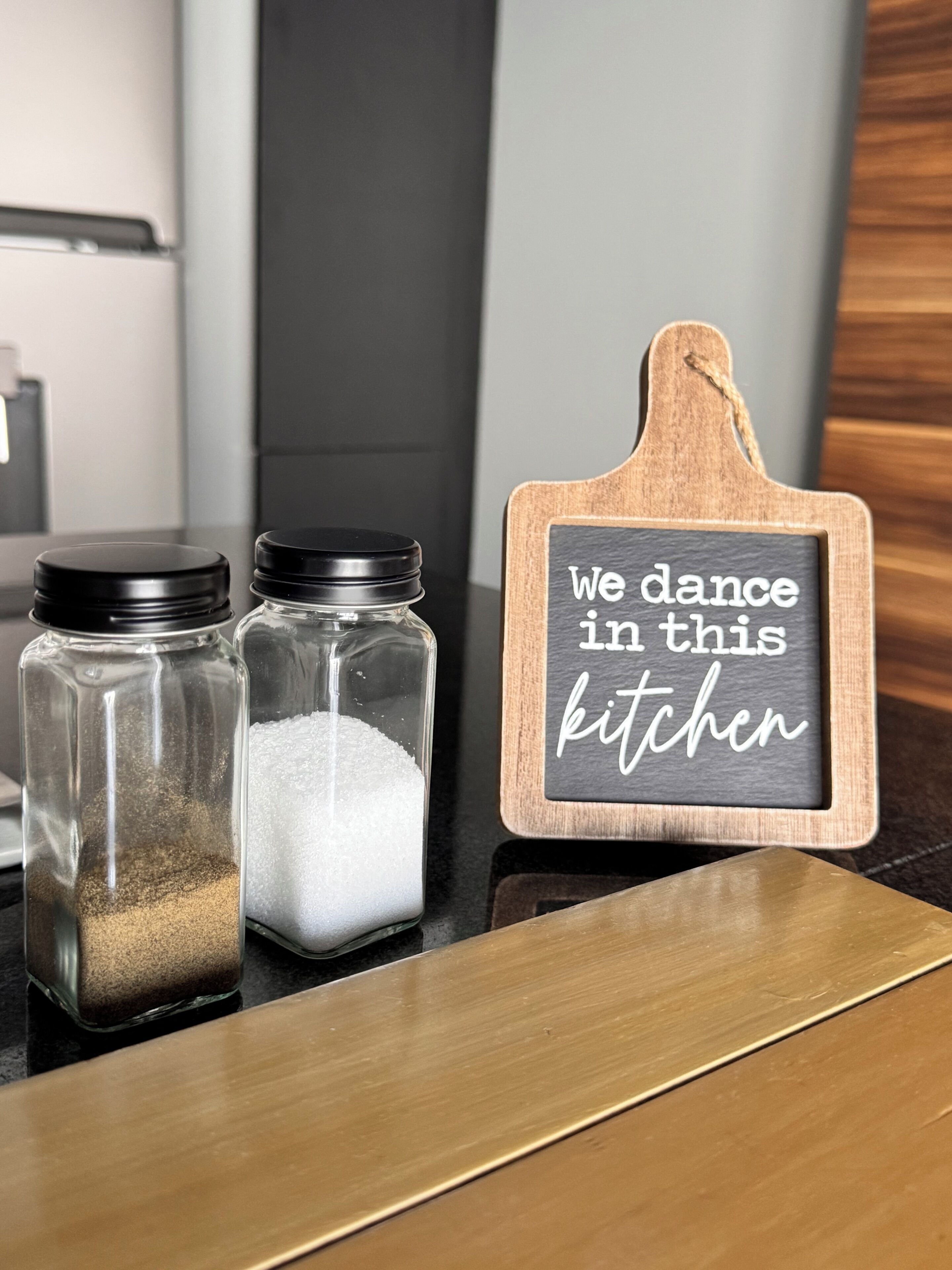 Zwei Gläser mit Deckeln auf einer Küchenarbeitsplatte, eines gefüllt mit einer bräunlichen Substanz, das andere mit weißer. Ein Holzschild mit der Aufschrift 'we dance in this kitchen' ist ebenfalls zu sehen.