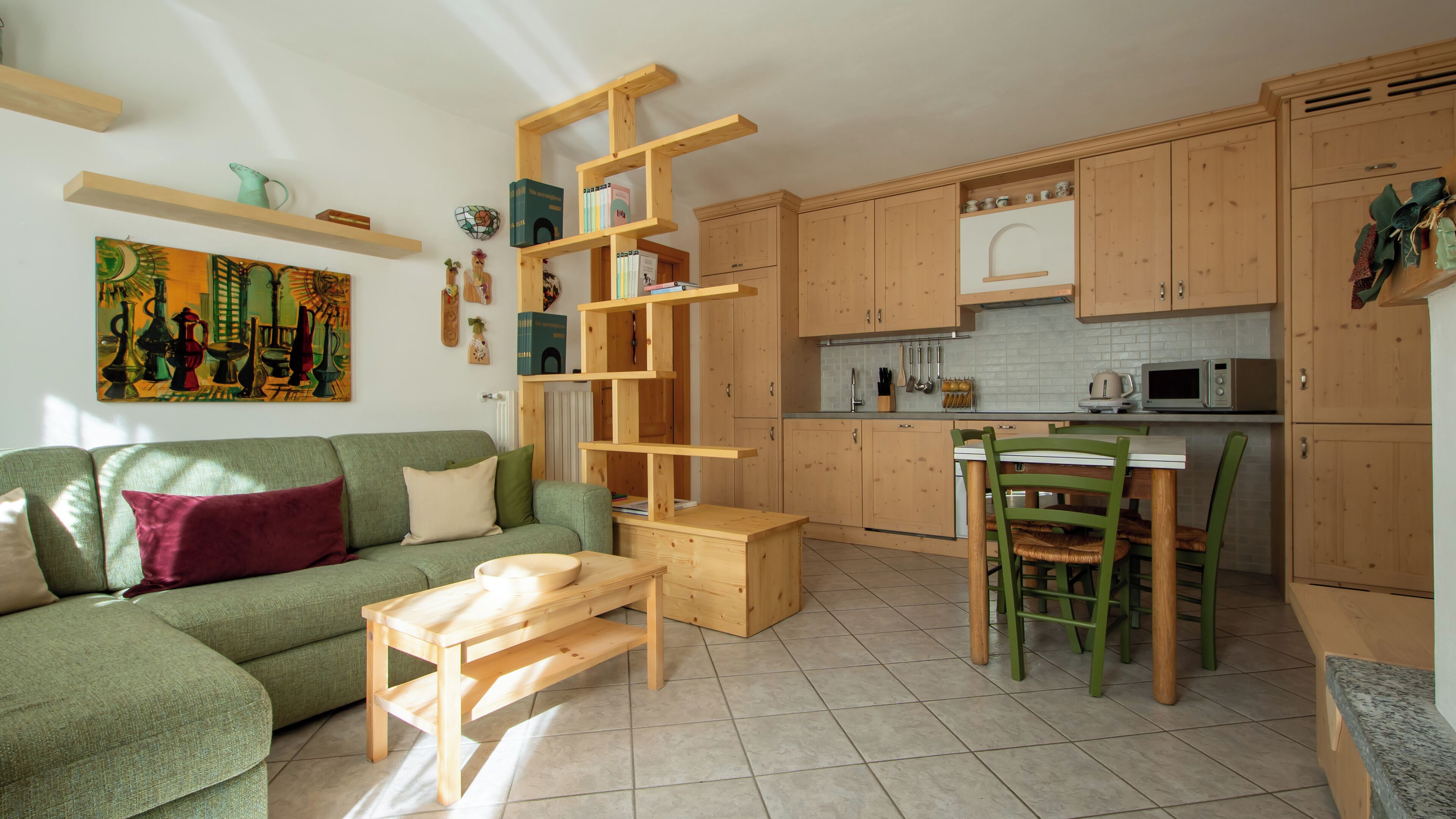 Ein gemütliches Wohnzimmer mit einem grünen Sofa, einem Holztisch und einem Bücherregal aus Holz. Die Küche verfügt über Holzschränke, eine Mikrowelle und ein Spülbecken.