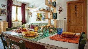 Spiseområde med et bord dækket til fire personer, med farvet glas og tallerkener. Der er en reol med bøger og pynteting, et vindue med gardiner, og en dør med en hjerteformet dekoration.