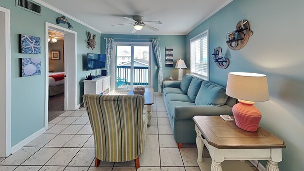 A living room with a ceiling fan, a sofa, a striped chair, a wooden table with a lamp, and a TV. - Pam's Paradise (Carolina Beach)