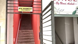 Una scala conduce a una stanza con la scritta "AFIE ROOMSTAY (SASA ROOMSTAY)", dotata di ringhiera in metallo e scherma a rete sulla destra.