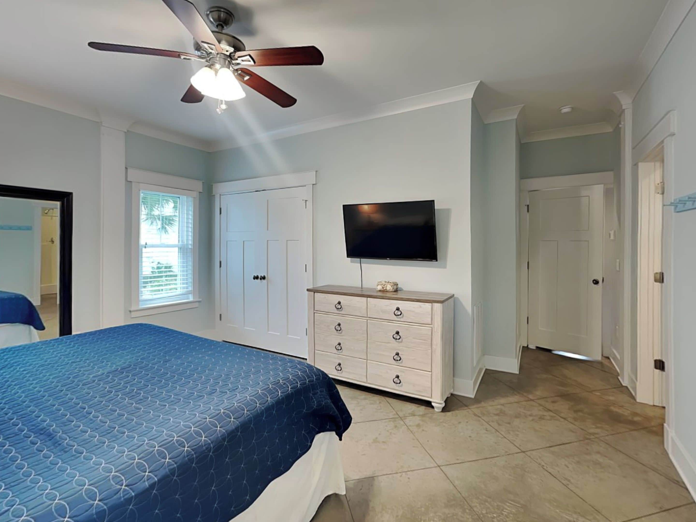 A bedroom with a bed, a ceiling fan, a TV, a dresser, and a door leading to another room.