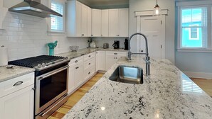 A modern kitchen with white cabinets, a stainless steel range hood, a built-in oven, a granite countertop, a sink with a flexible faucet, and pendant lighting. - Seahorse by the Sea (Kure Beach)