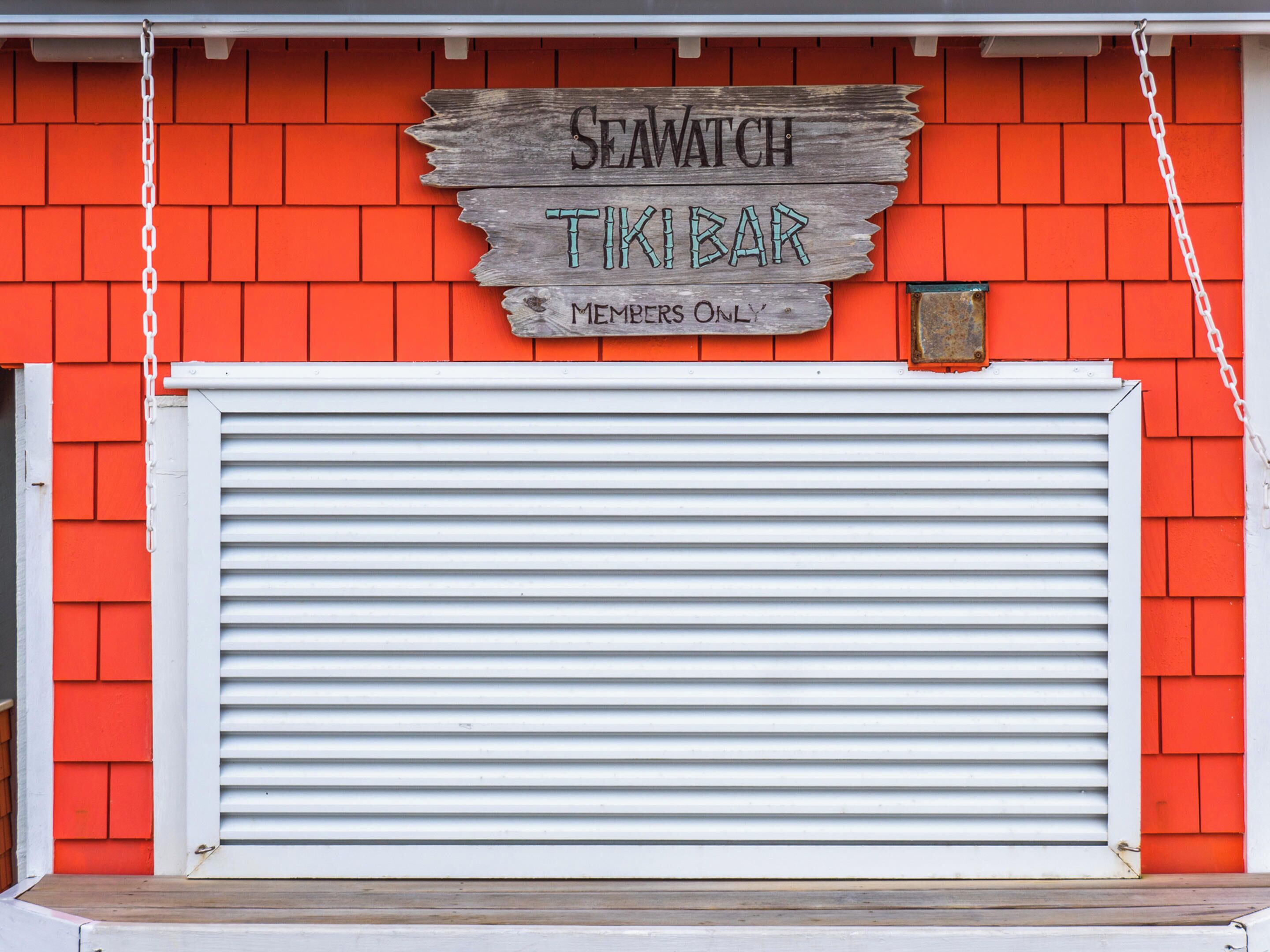 A wooden sign with 'Seawatch Tiki Bar' and 'Members Only' text, mounted on a red brick wall with a white shutter door below.