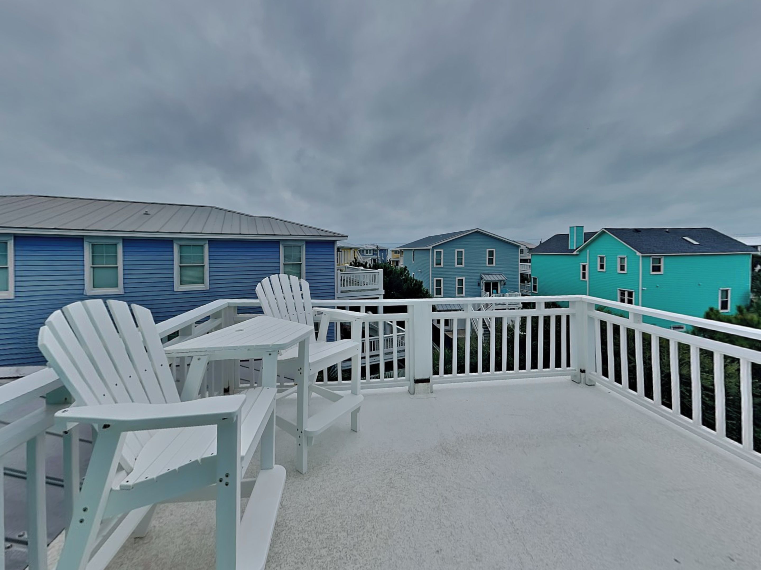 A covered outdoor deck with white Adirondack chairs and a table, overlooking a row of colorful houses.