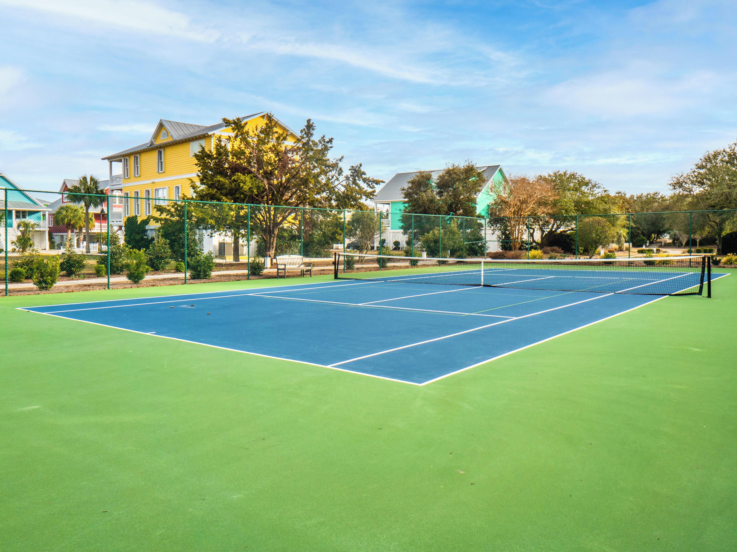 An outdoor tennis court with a green surface, surrounded by a fence, and adjacent to a yellow building with a green roof.