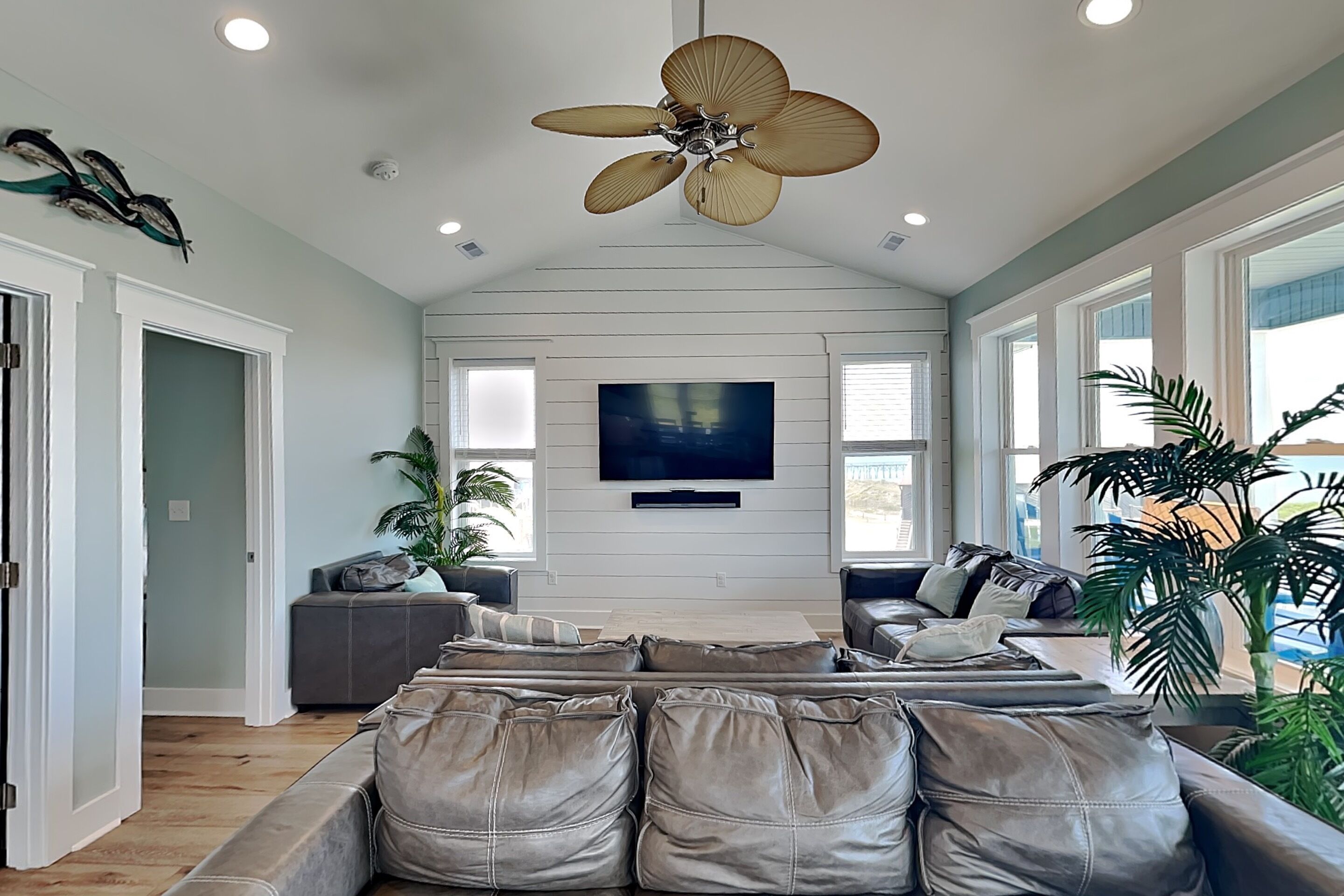 Un salón amplio con un sofá grande de cuero, un ventilador de techo, un televisor de pantalla plana y ventanas grandes.