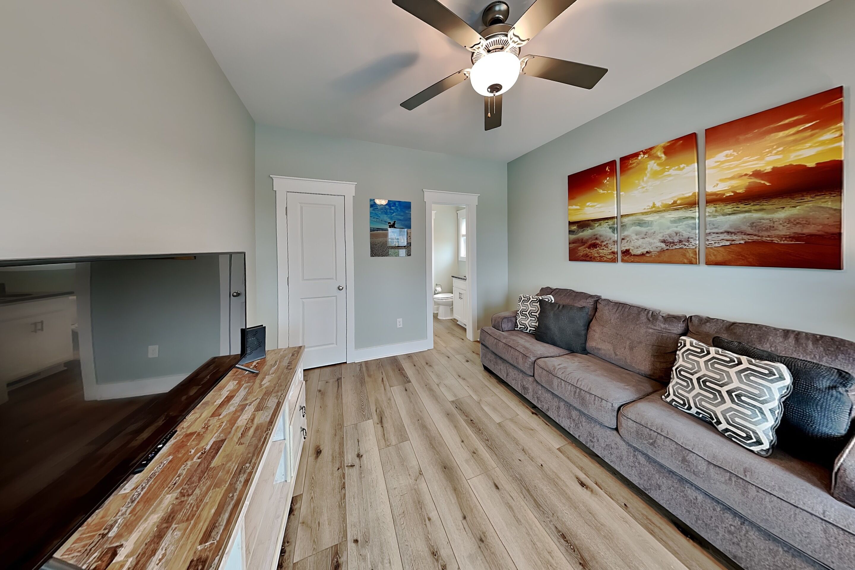 A modern living room with a wooden coffee table, a grey sofa with patterned cushions, a ceiling fan, and a large wall art depicting a sunset over the ocean.