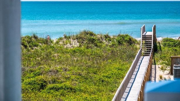 A wooden boardwalk leading to a beach with clear blue water and green vegetation. - Good Tide-ings (Sneads Ferry)