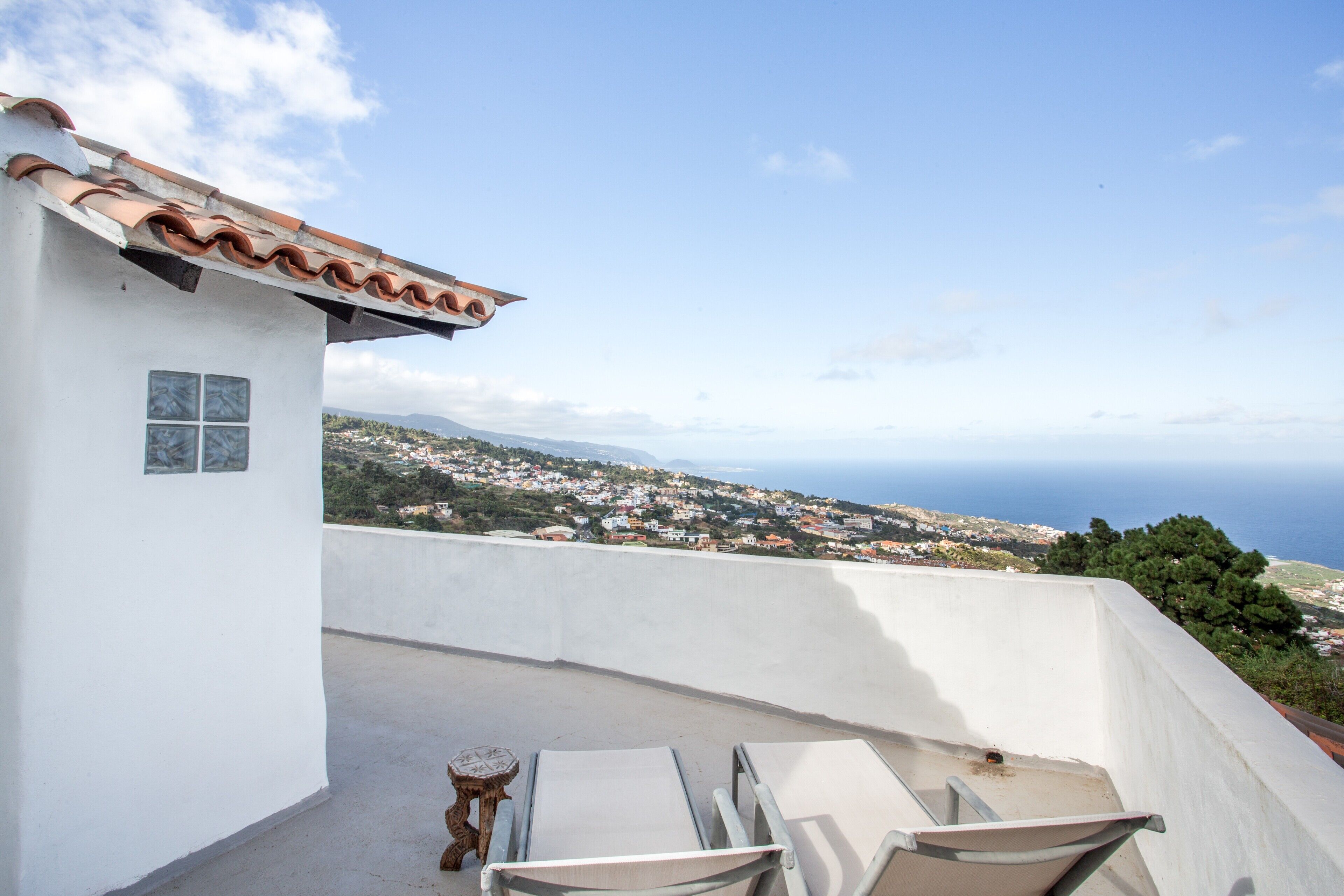 A white building with a tiled roof, a small table, and two chairs on a balcony overlooking a coastal town.