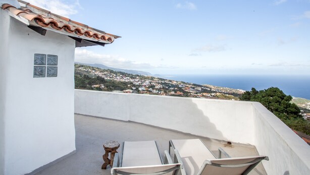 Un bâtiment blanc avec un toit recouvert de tuiles, une petite table et deux chaises sur un balcon donnant sur une ville côtière.