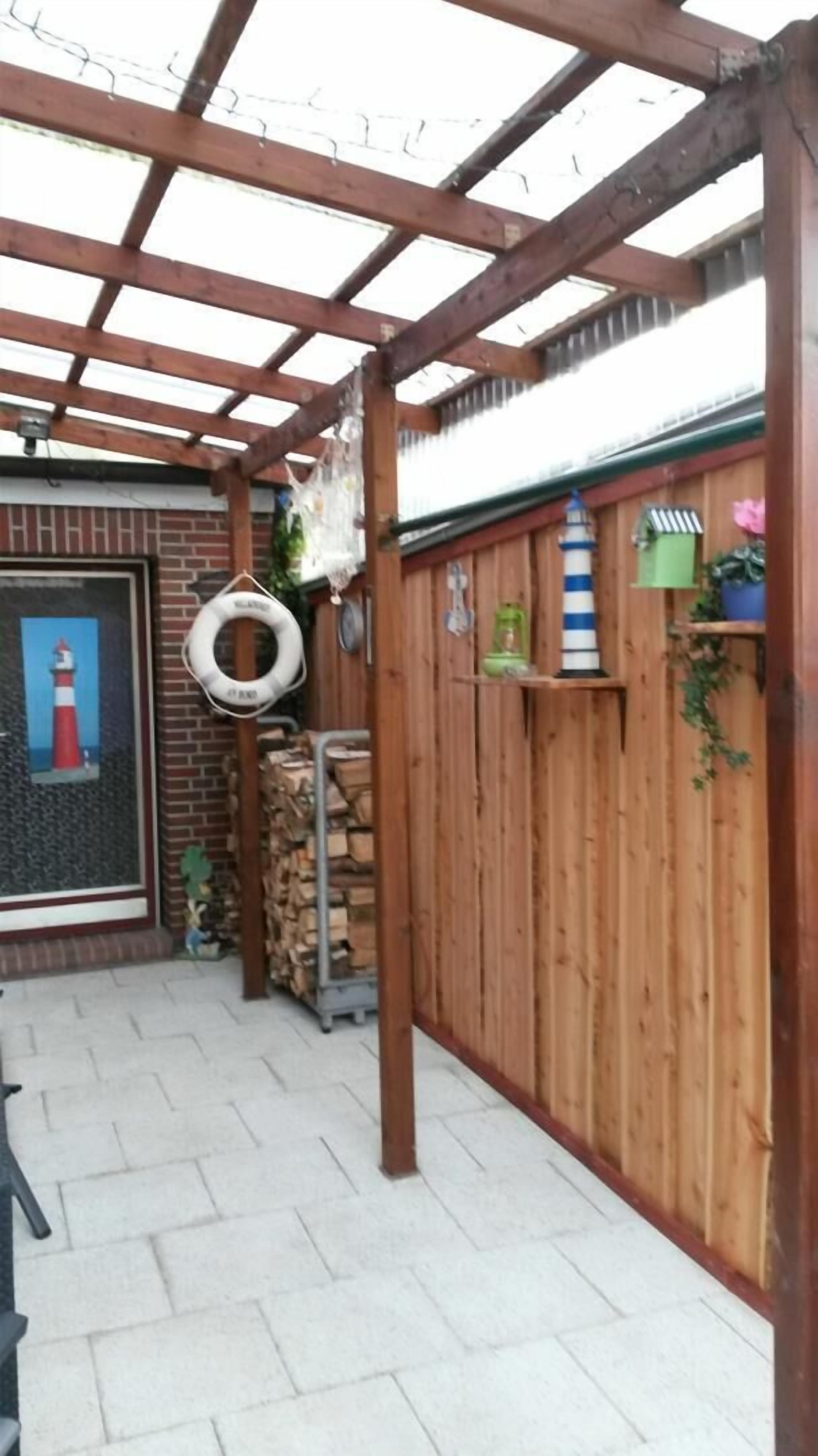 En pergola av trä med ett tak av metall, en mur av tegel med ett fönster och ett staket av trä med en grön växt.