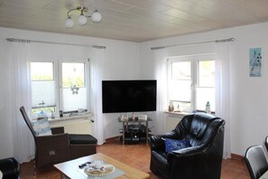 Una sala de estar con un televisor de pantalla plana, un sofá de cuero negro, una silla de mimbre y una mesa de comedor.