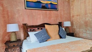 Un dormitorio con cama, dos mesitas de noche con lámparas, un cuadro enmarcado en la pared y una colcha estampada.