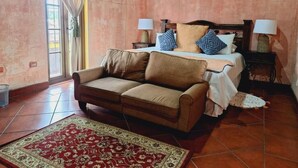 Un dormitorio con cama, sofá, mesita de noche, lámpara, ventana con cortinas y un cuadro en la pared.