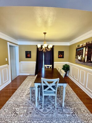 A dining room with a dark wooden table, white chairs, a chandelier, and patterned rugs.