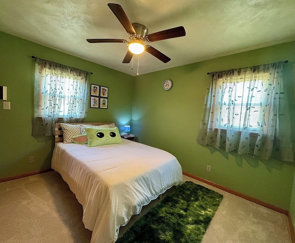 A bedroom with a bed, a ceiling fan, two windows with curtains, a wall clock, and a small table with a lamp.