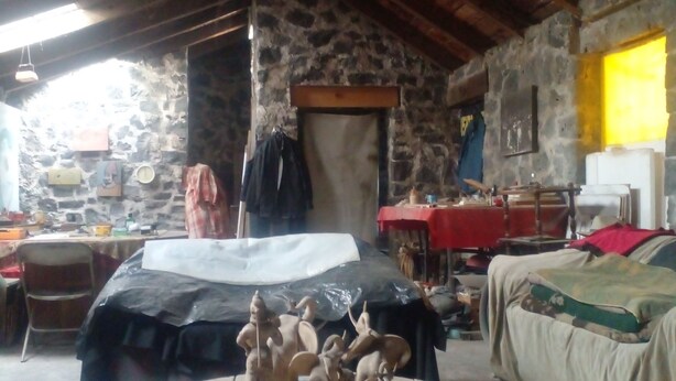 Quarto rústico com parede de pedra, teto de madeira e uma mesa redonda com miniaturas. Há uma cama com cobertor vermelho e uma mesa com diversos objetos.