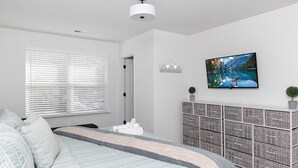 Een slaapkamer met een bed, een dressoir, een televisie en een raam met jaloezieën.