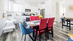 Een moderne keuken met witte kasten, een magnetron en een koelkast. Er is een eethoek met een tafel en stoelen, en een tafelvoetbal in de hoek.